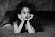 Home A thoughtful young woman with curly hair lies on a bed in a black and white portrait. Moody and introspective.